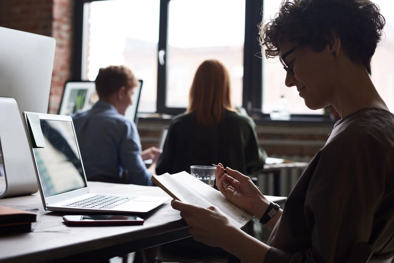 Woman with curly hair and glasses reading a notebook at a desk with a laptop and smartphone, two people are blurred in the background.
